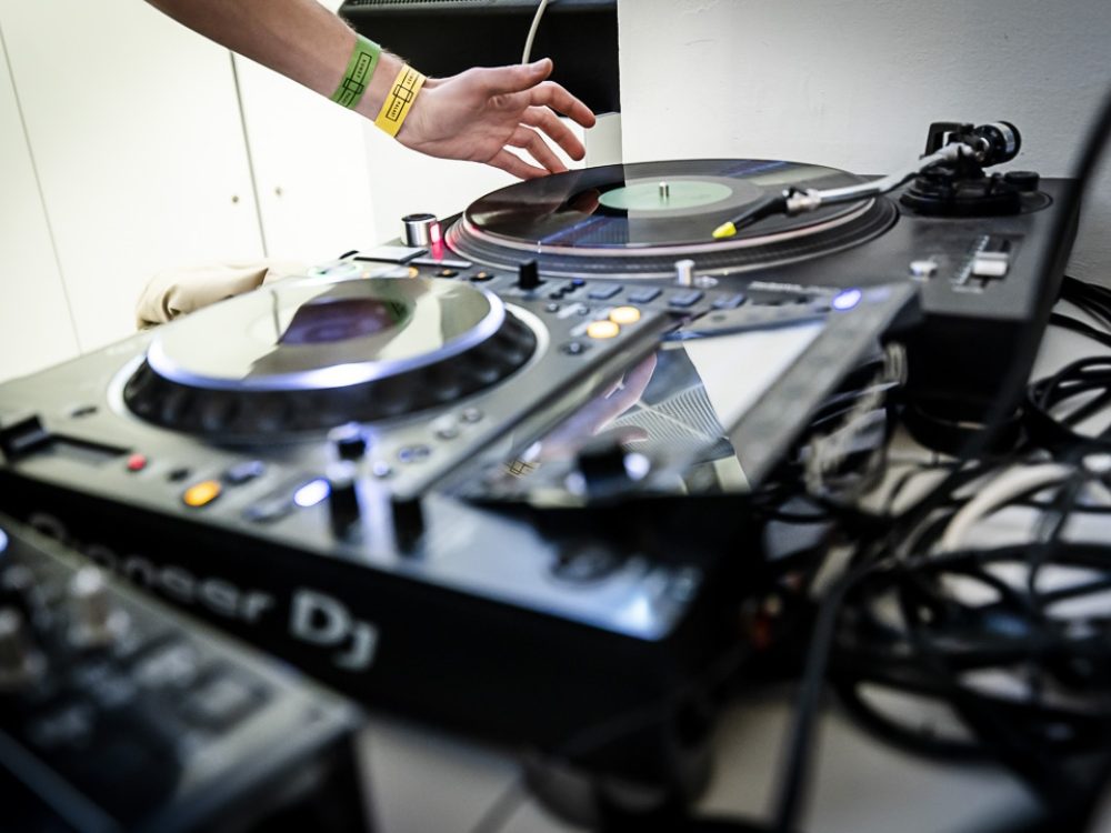 Nahaufnahme eines DJs, der mit der Hand einen Plattenspieler und ein Mischpult einstellt, mit verschiedenen Geräten auf einem Tisch.