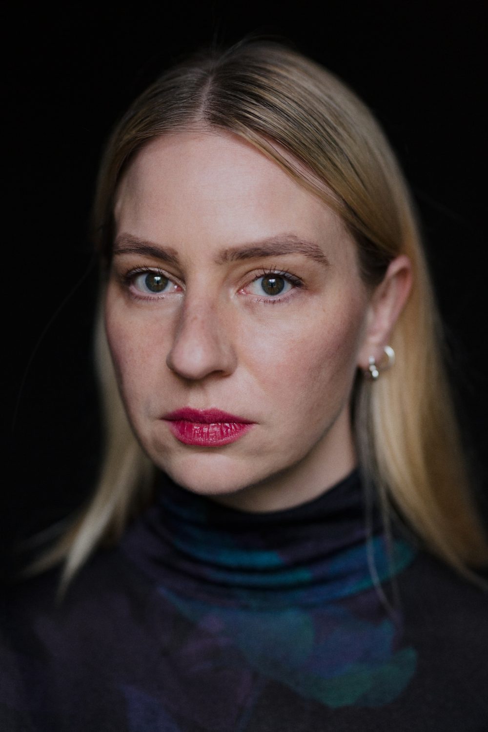 Eine Frau mit blondem Haar, die einen dunklen Rollkragenpullover trägt, blickt ernst in die Kamera vor einem schwarzen Hintergrund.