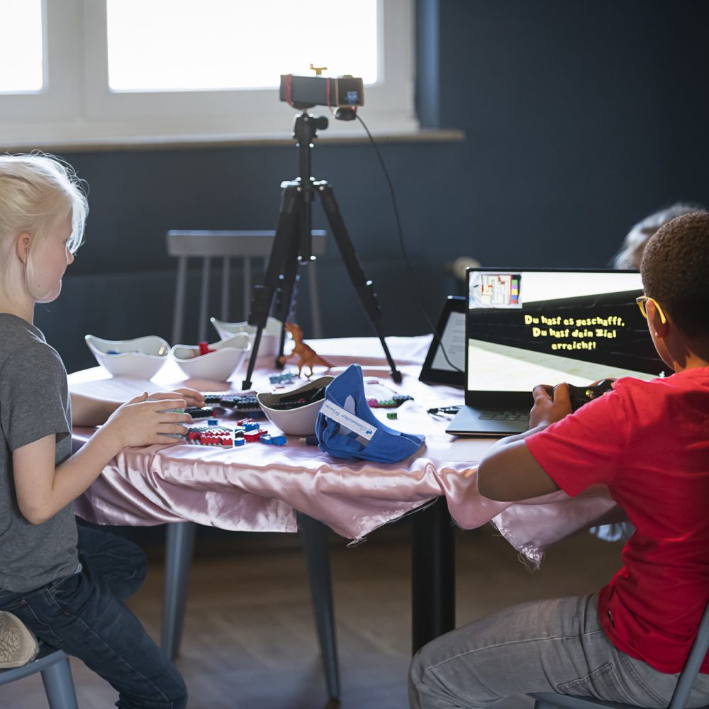 Zwei Kinder sitzen an einem Tisch auf dem ein Laptop mit geöffnetem Spiel steht sowie Legosteine und ein Stativ mit Kamera sind