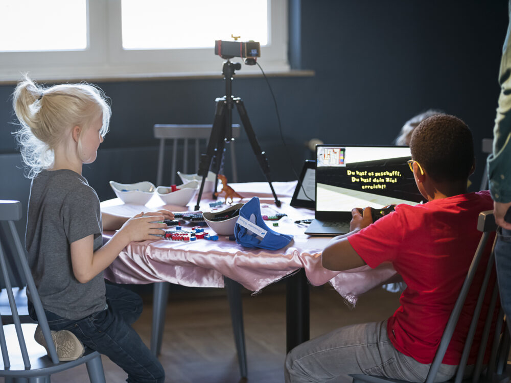 Two children sit at a table with a laptop with an open game, Lego bricks and a tripod with a camera