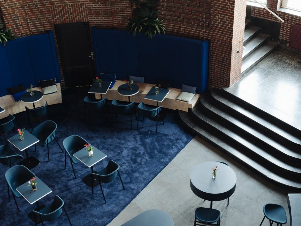 Gastronomie Pong mit mehreren blauen, mit Blumen dekorierten Tischen und blauen Stühlen auf einem blauen Teppich vor roten Backsteinwand