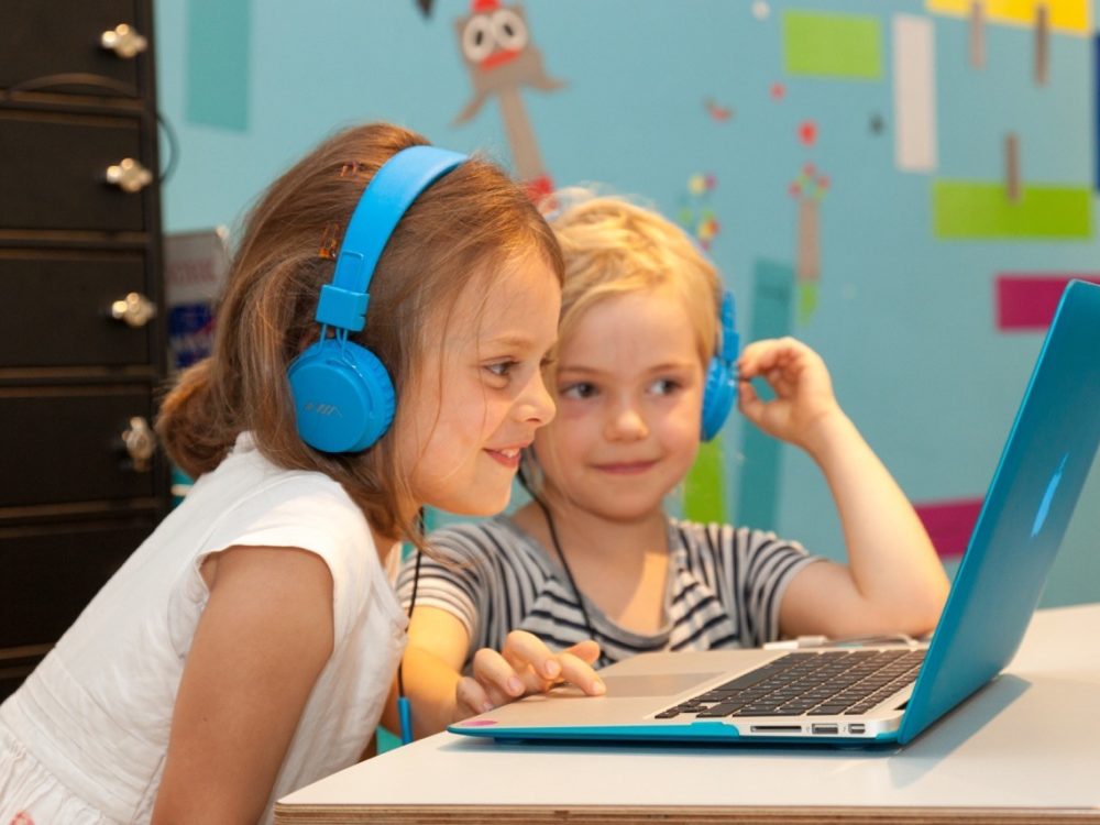Zwei kinder sitzen vor einem Laptop mit blauer Schutzhülle und haben blaue Kopfhörer auf vor einer bunten Wand im Hintergrund
