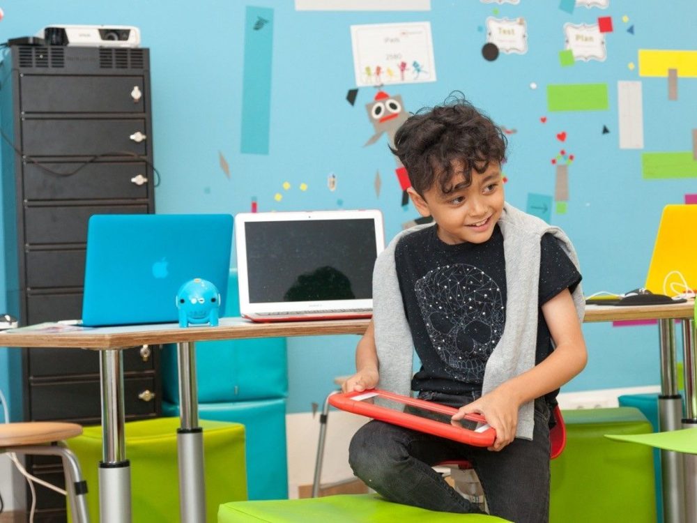Ein kleiner Junge kniet mit einem Bein auf einem Hocker und hat ein rotes Tablet in der Hand vor einem Tisch mit Laptops und einer bunten Wand