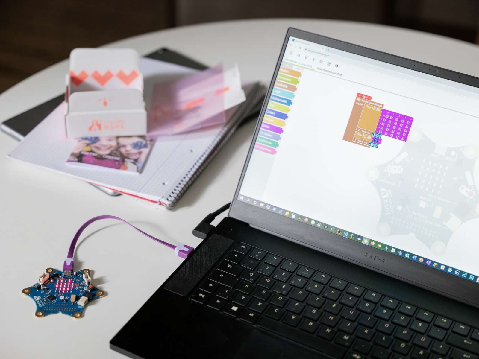 Ein aufgeklappter Laptop mit geöffnetem Codierungsprogramm, mit dem eine Platine verbunden ist steht auf einem weißen Tisch mit Unterlagen