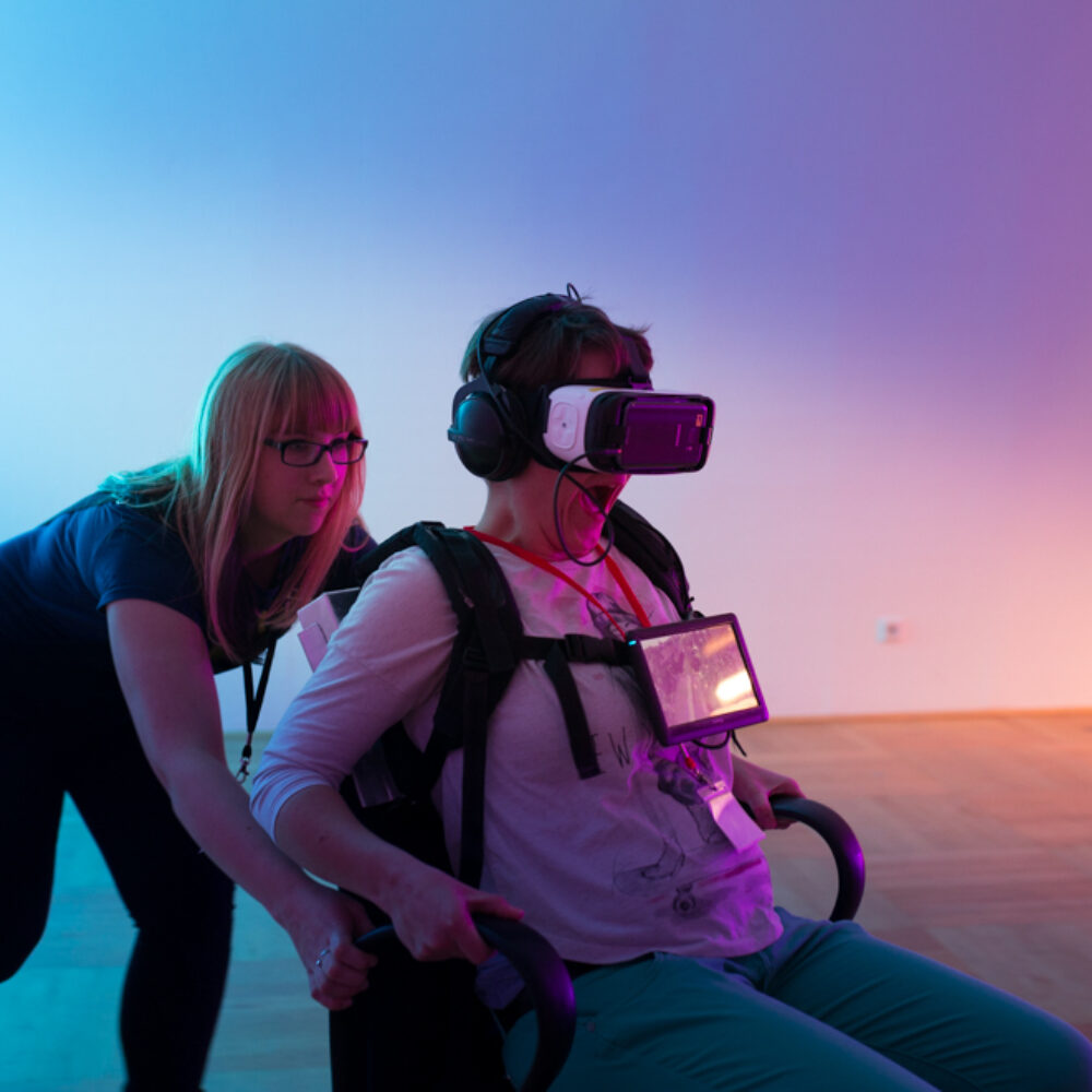 Eine Person sitzt mit einer VR Brille auf dem Kopf auf einem Bürostuhl, an dem sie sich festzuhalten versucht und wird von einer jungen Frau geschoben