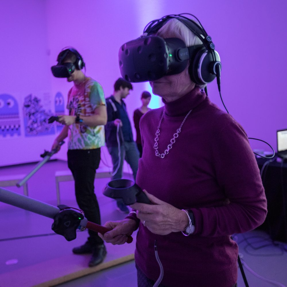 Ältere Frau steht in lila beleuchtetem Raum und hat VR Brille auf dem Kopf sowie einen Controller und ein elektronisches Schwert in der Hand
