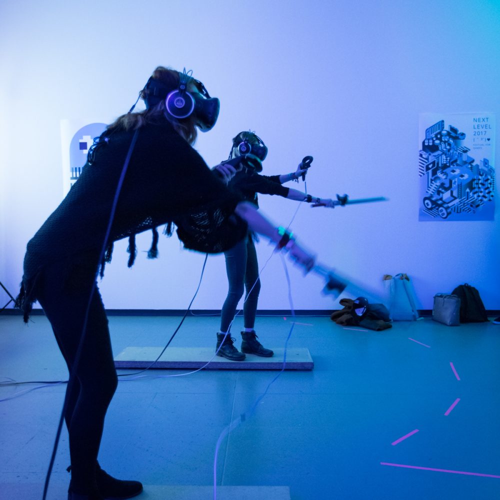 Zwei Personen spielen in einem blau beleuchteten Raum mit VR Brillen auf dem Kopf und Controllern in der Hand