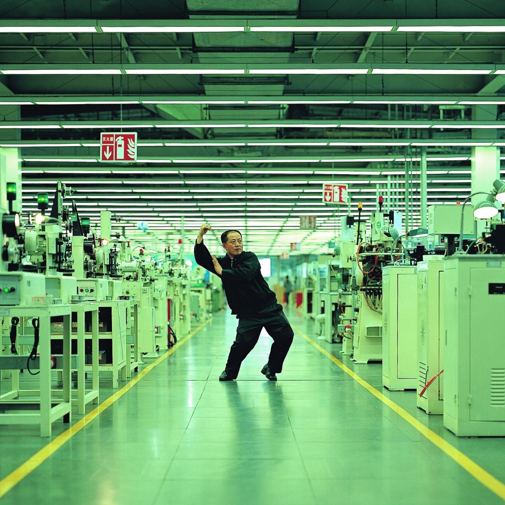 Eine Person posiert energisch inmitten einer hell erleuchteten, modernen Fabrik voller Industriemaschinen.