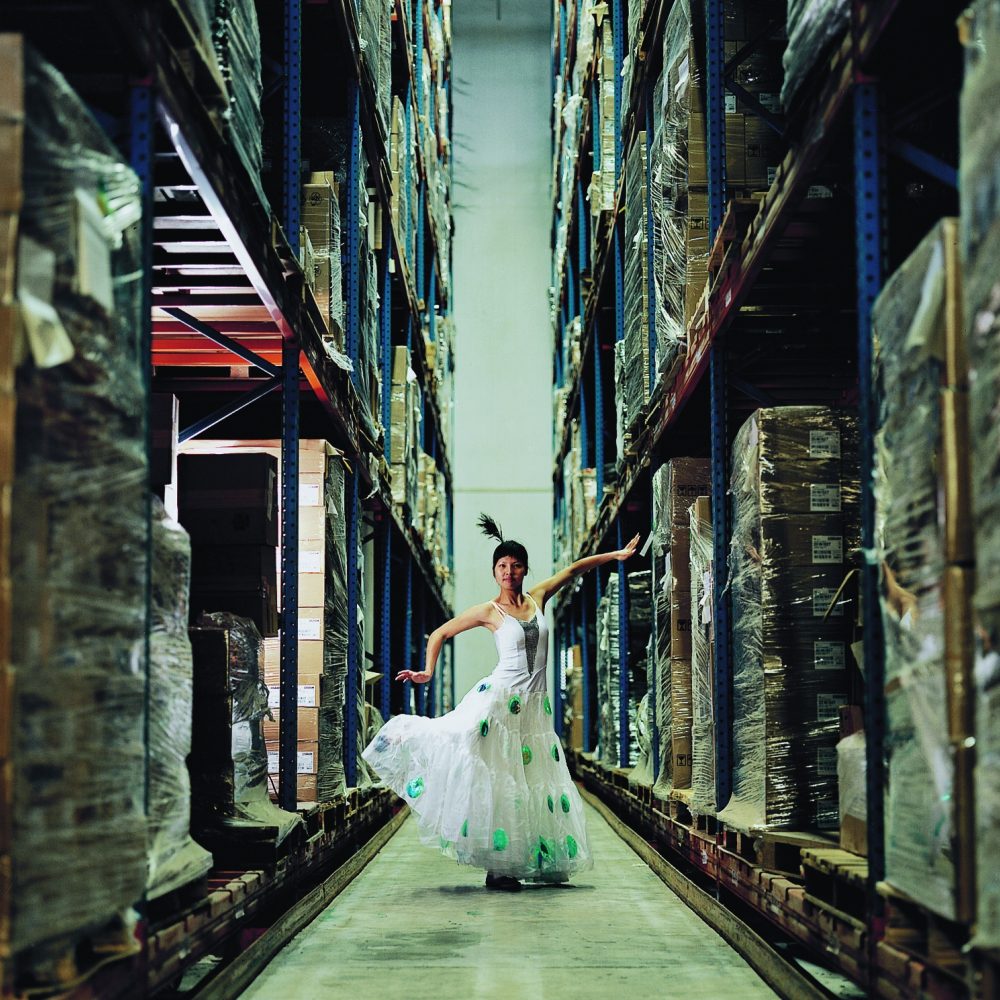 Eine Frau in einem weißen Kleid tanzt zwischen hohen Kistenregalen in einem Lagerhaus.