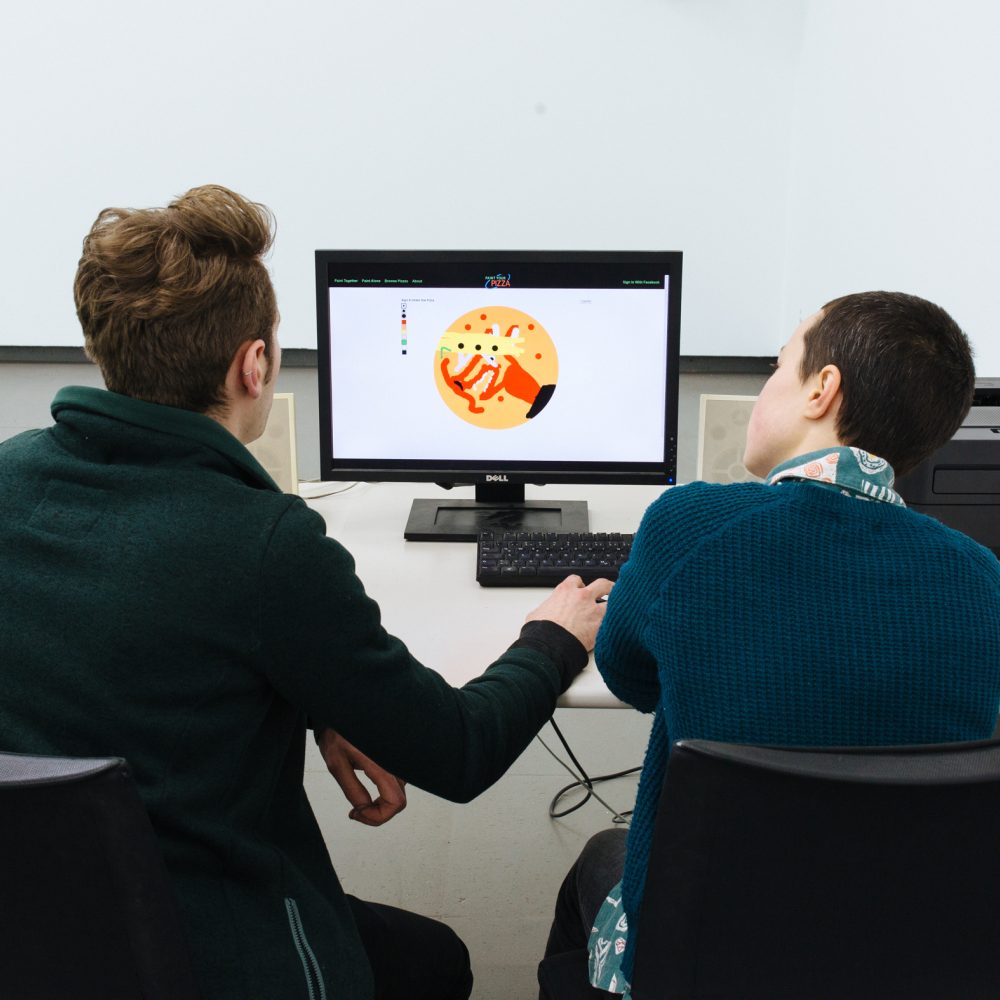 Two persons sit in front of a computer in the exhibition Pizza is God and paint a pizza with a program
