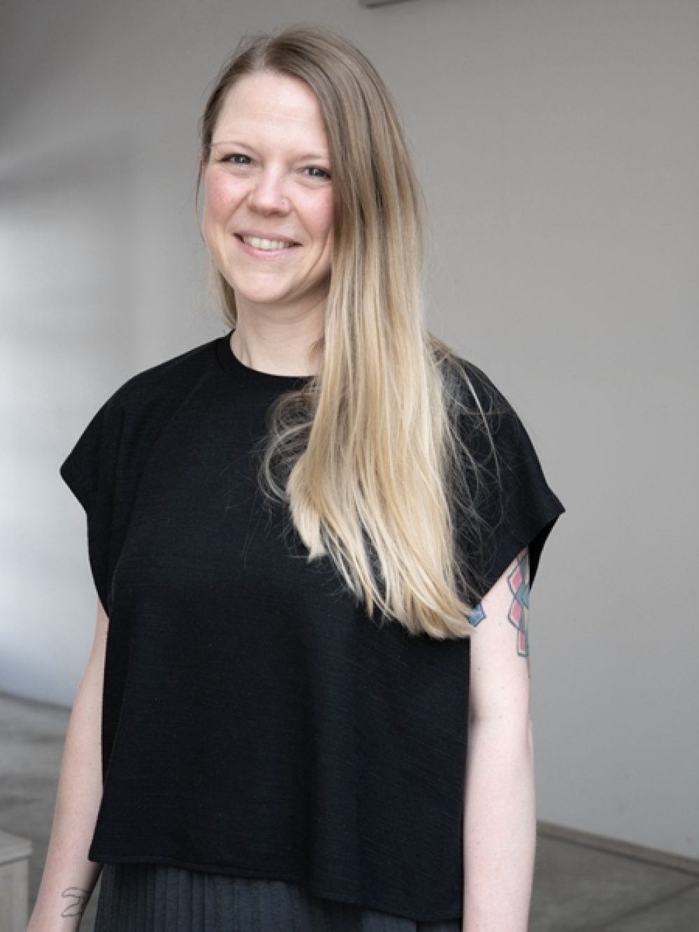 Eine lächelnde Frau mit langen blonden Haaren, die ein schwarzes Oberteil trägt, steht in einem Haus und hat eine Tätowierung auf ihrem linken Arm.