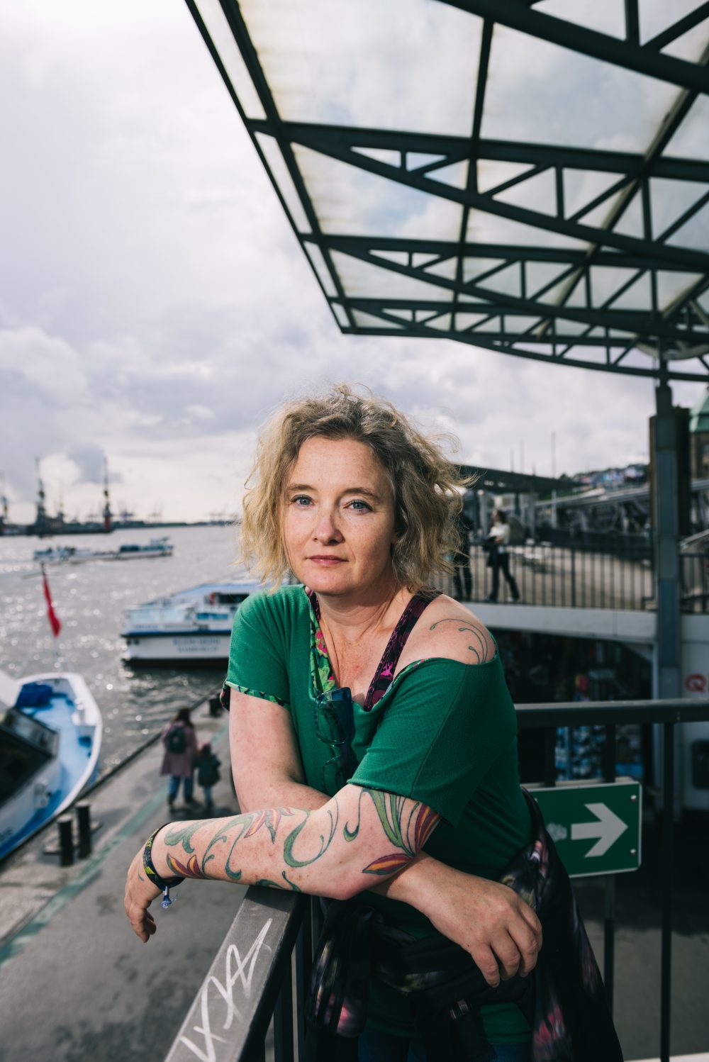Eine Frau mit lockigem blondem Haar und tätowierten Armen lehnt an einem Geländer an einem Flussufer mit Booten und bedecktem Himmel.