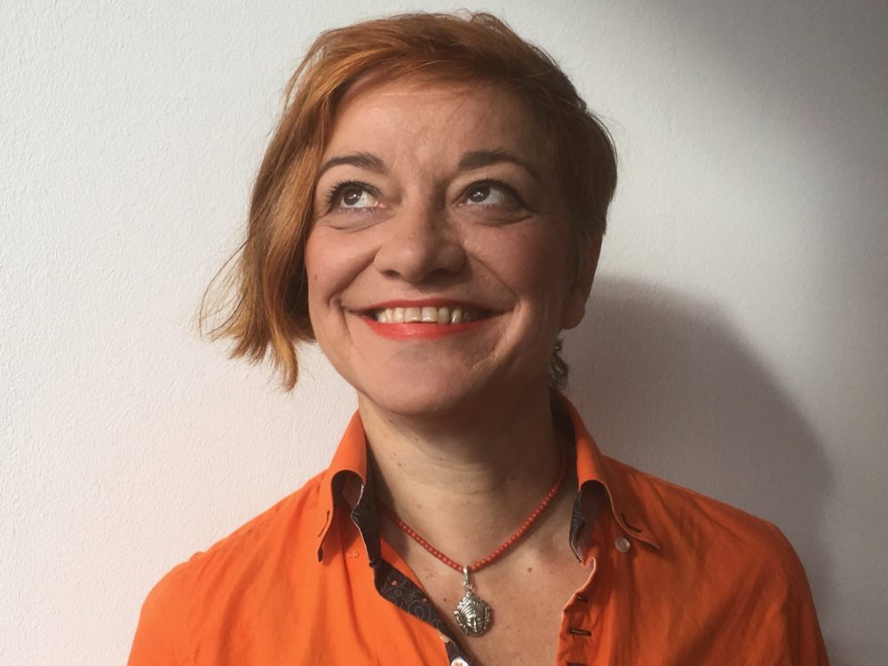 Eine lächelnde Frau mit kurzen roten Haaren in einem orangefarbenen Hemd blickt an einer schlichten weißen Wand nach oben.