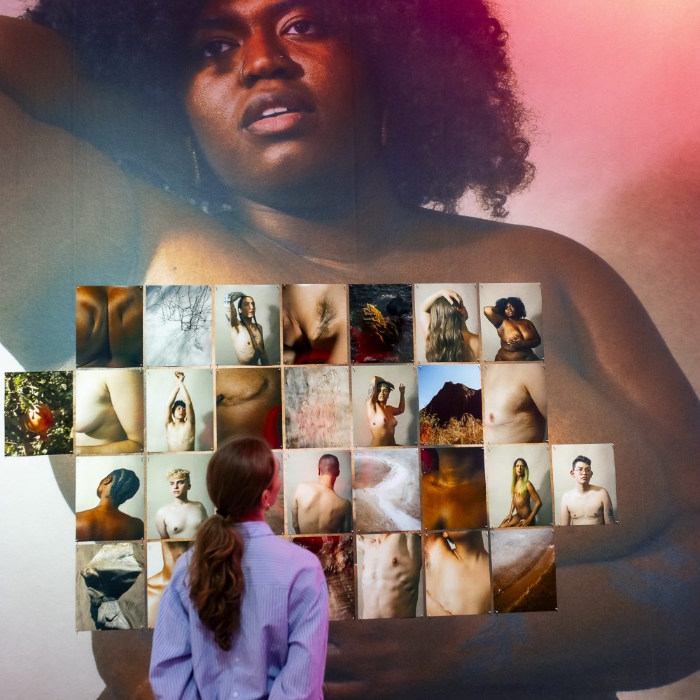 Eine Person betrachtet eine Wand mit verschiedenen Aktfotografien vor dem Hintergrund eines großen Porträts einer Frau mit einem Afro.