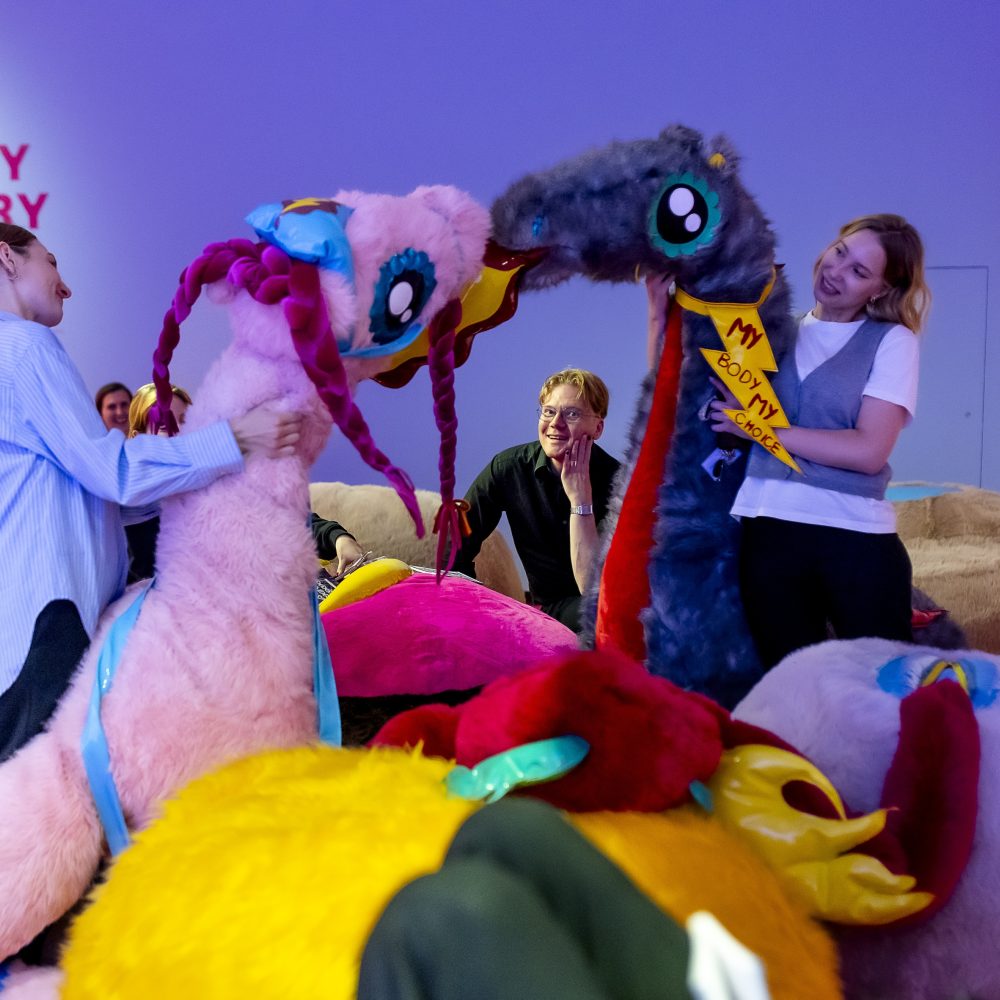 Zwei Frauen lachen, während sie spielerisch große, bunte Plüschtiere in einem lebhaften, skurrilen Raum mit der Aufschrift Fluffy Library halten.