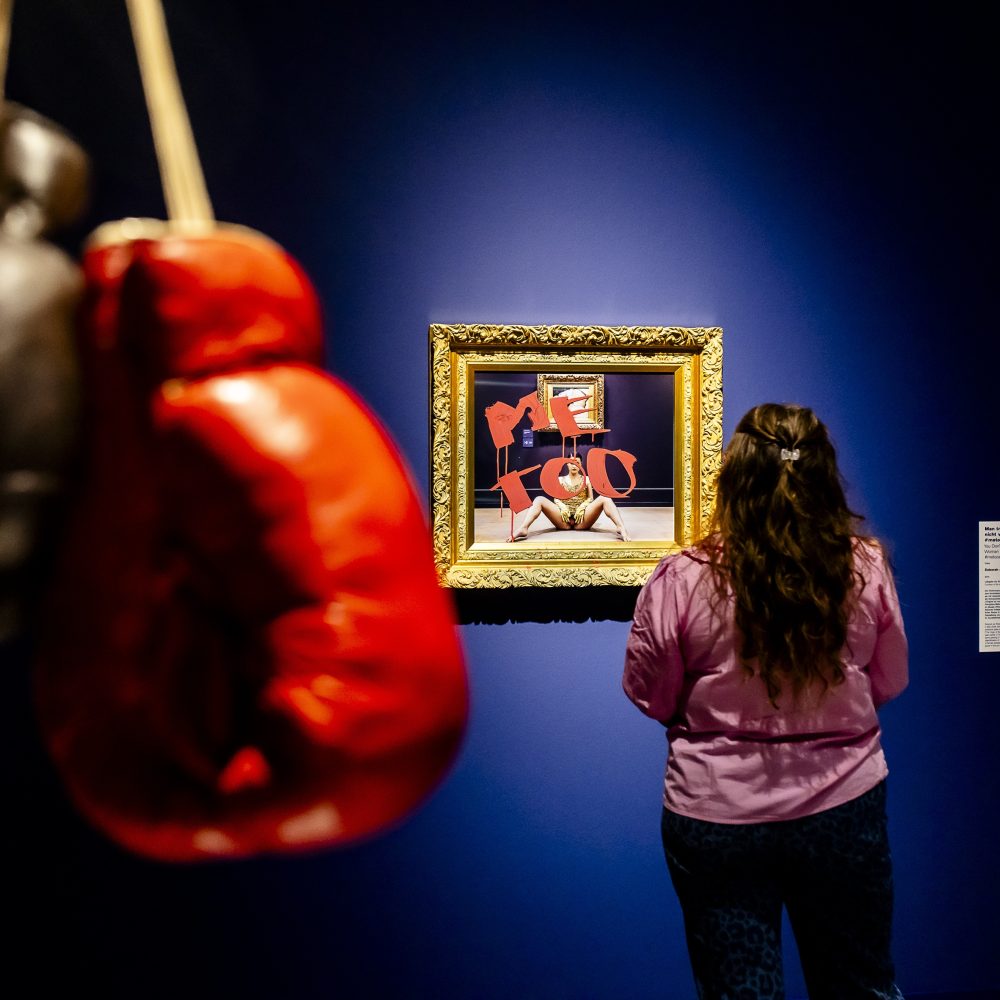 Eine Frau betrachtet ein gerahmtes Kunstwerk in einer Galerie; im Vordergrund hängen rote und schwarze Boxhandschuhe.