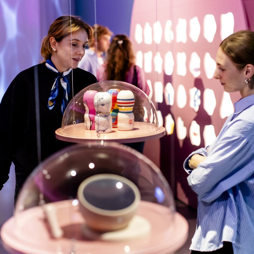 Zwei Personen betrachten Regale mit bunten Gegenständen unter Glaskuppeln in einem modernen, hell erleuchteten Ausstellungsraum.