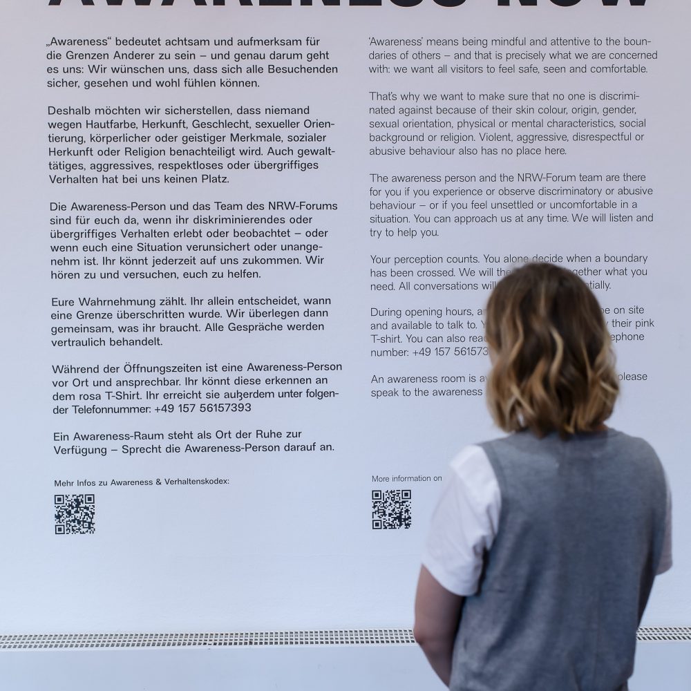 Eine Person steht vor einer Wand mit einem großen Text mit der Überschrift AWARENESS NOW in deutscher und englischer Sprache.