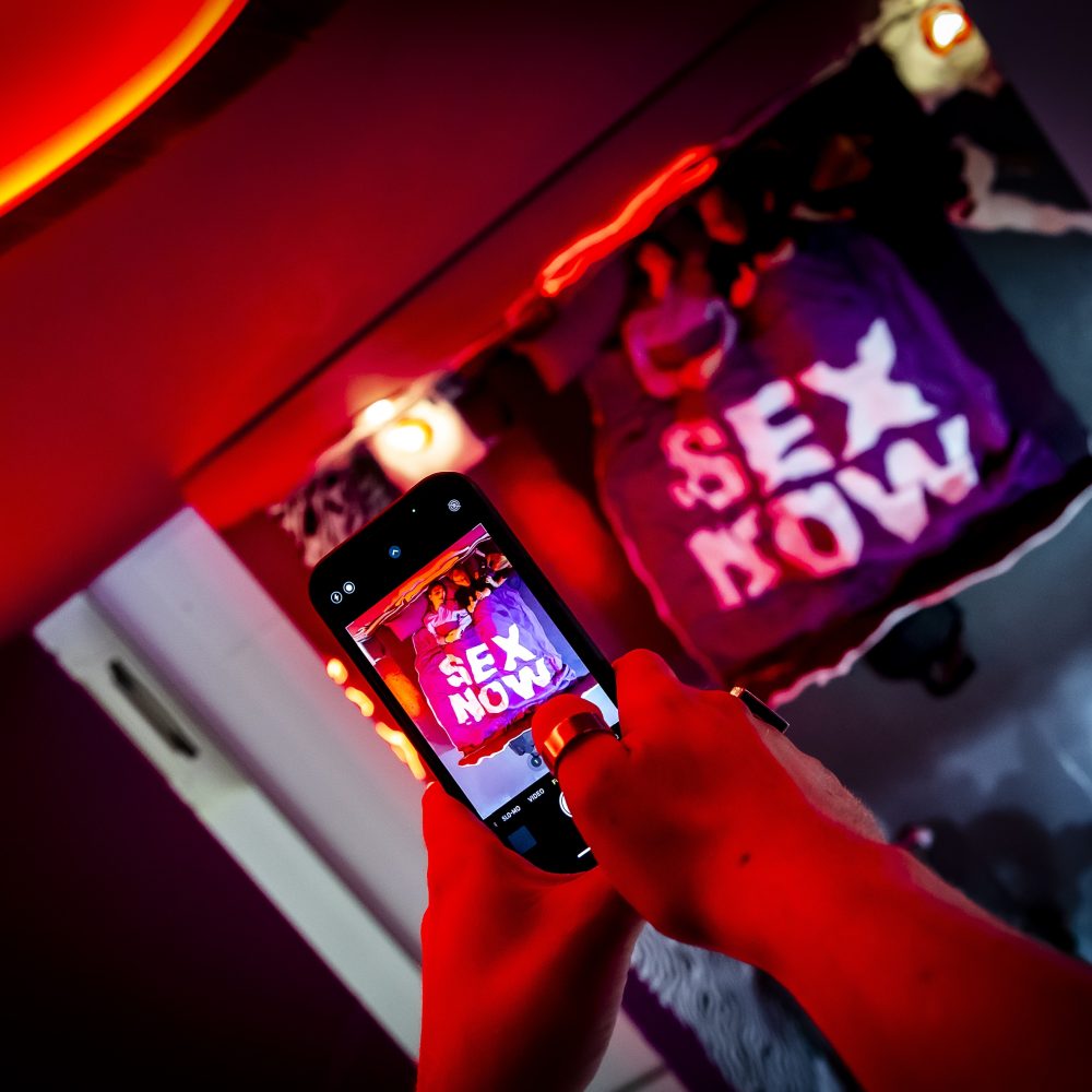 Eine Person fotografiert ein Hemd mit der Aufschrift "SEX NOW" in einem von roten und violetten Lichtern beleuchteten Raum.