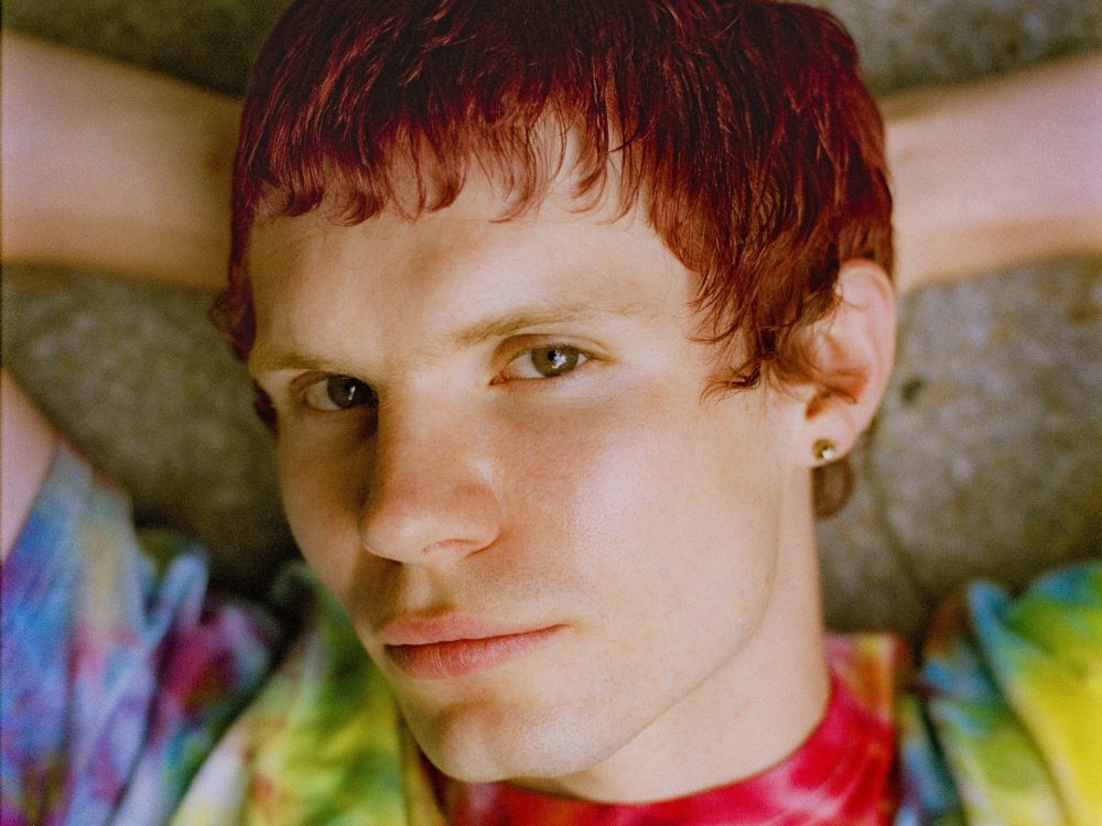 Eine Person mit kurzen roten Haaren und einem Batikhemd liegt auf dem Rücken, die Arme hinter dem Kopf und schaut in die Kamera.