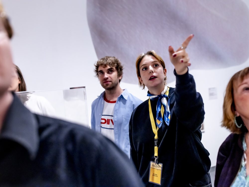 Eine Frau mit einem Schlüsselband zeigt auf eine Gruppe von Personen, die sich in einem Haus befinden und in die angegebene Richtung schauen.