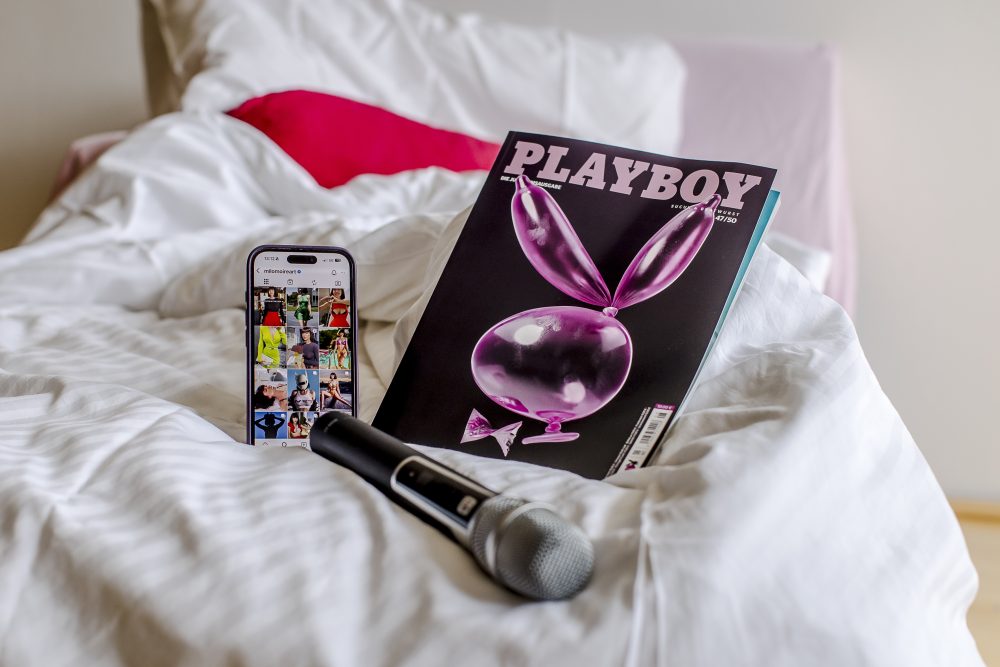 Ein Playboy-Magazin, ein Smartphone und ein Mikrofon liegen auf einem weißen Bett mit rosa Kissen im Hintergrund.