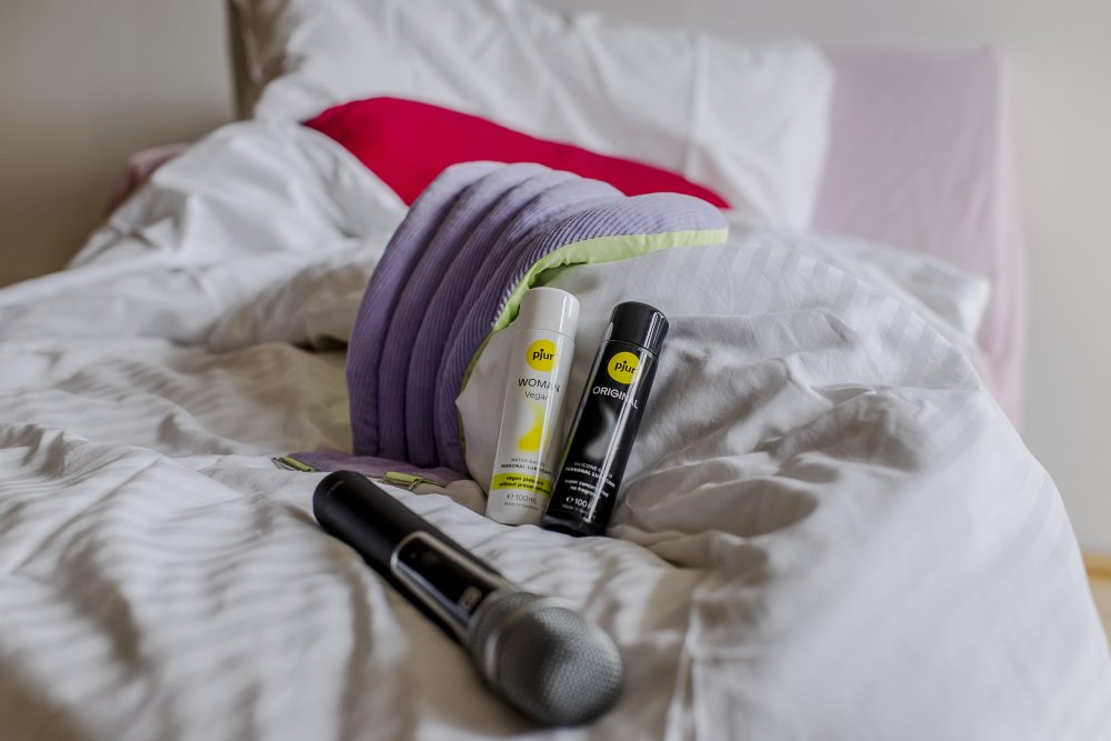 Ein Mikrofon und zwei Flaschen mit der Aufschrift pjur liegen auf einem zerknitterten Bett mit weißen Laken und bunten Kissen im Hintergrund.