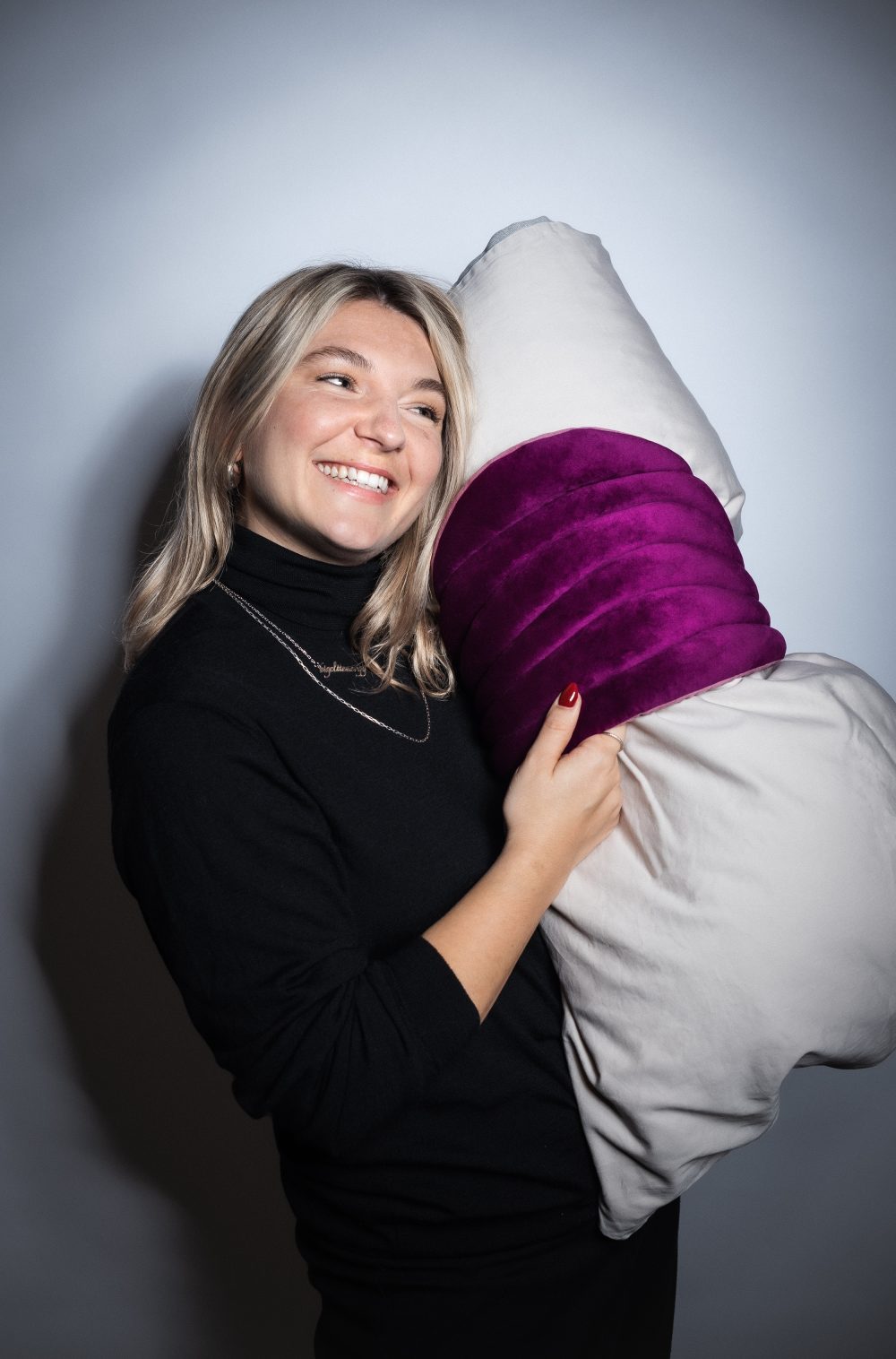 Lächelnde Frau in einem schwarzen Rollkragenpullover, die ein großes Kissen mit einem lila Streifen vor einem schlichten Hintergrund hält.