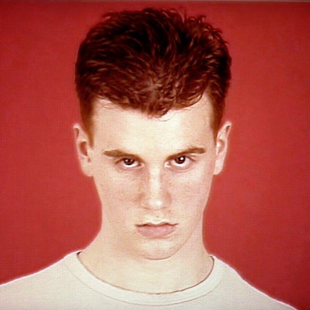 Young man with red hair and a whit t-shirt in front of a red background looking into the camera