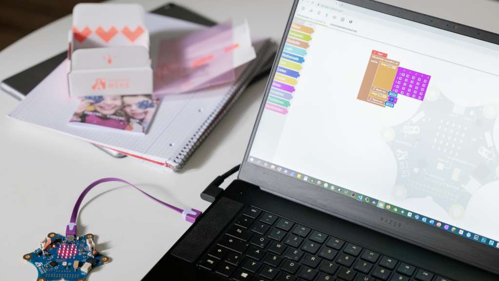 Ein aufgeklappter Laptop mit geöffnetem Codierungsprogramm, mit dem eine Platine verbunden ist steht auf einem weißen Tisch mit Unterlagen