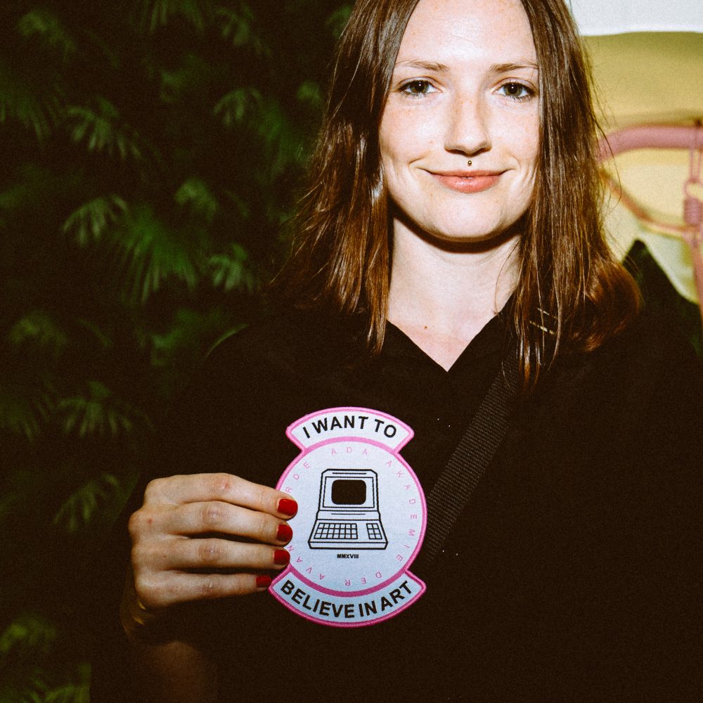 Eine junge Frau hält ein Badge vor sich auf dem I want to belive in art steht und ein Computer abgebildet ist