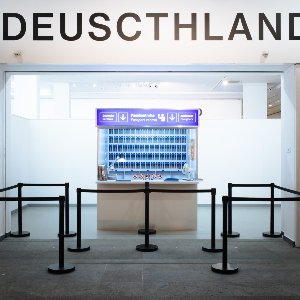 Ein Tresen mit blauen Tabletts unter einem falsch geschriebenen DEUSCTHLAND-Schild, davor bilden schwarze Absperrungen eine Warteschlange.