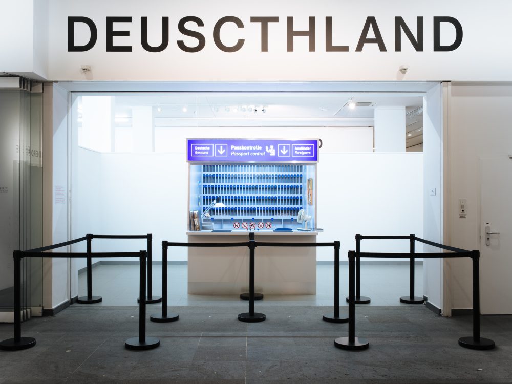 Ein Tresen mit blauen Tabletts unter einem falsch geschriebenen DEUSCTHLAND-Schild, davor bilden schwarze Absperrungen eine Warteschlange.