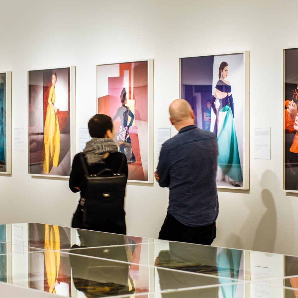 Zwei Personen schauen sich in der Ausstellung Horst Photographer of Style an der Wand hängende Fotografien an. Im Vorderungd stehen weiße Vitrienen