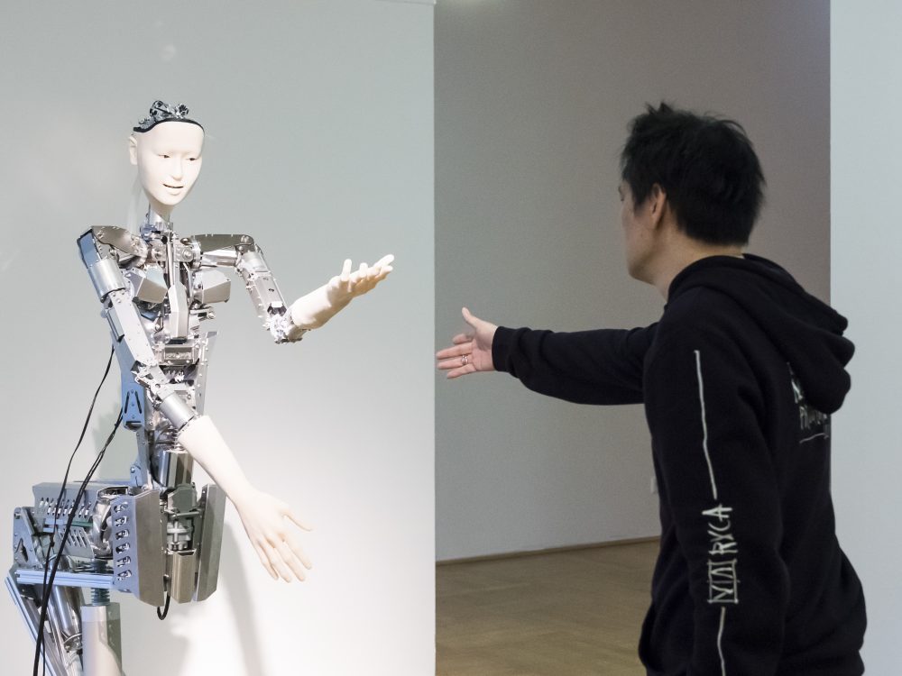 A black dressed man offers his hand to a robot in the exhibition Körperwende