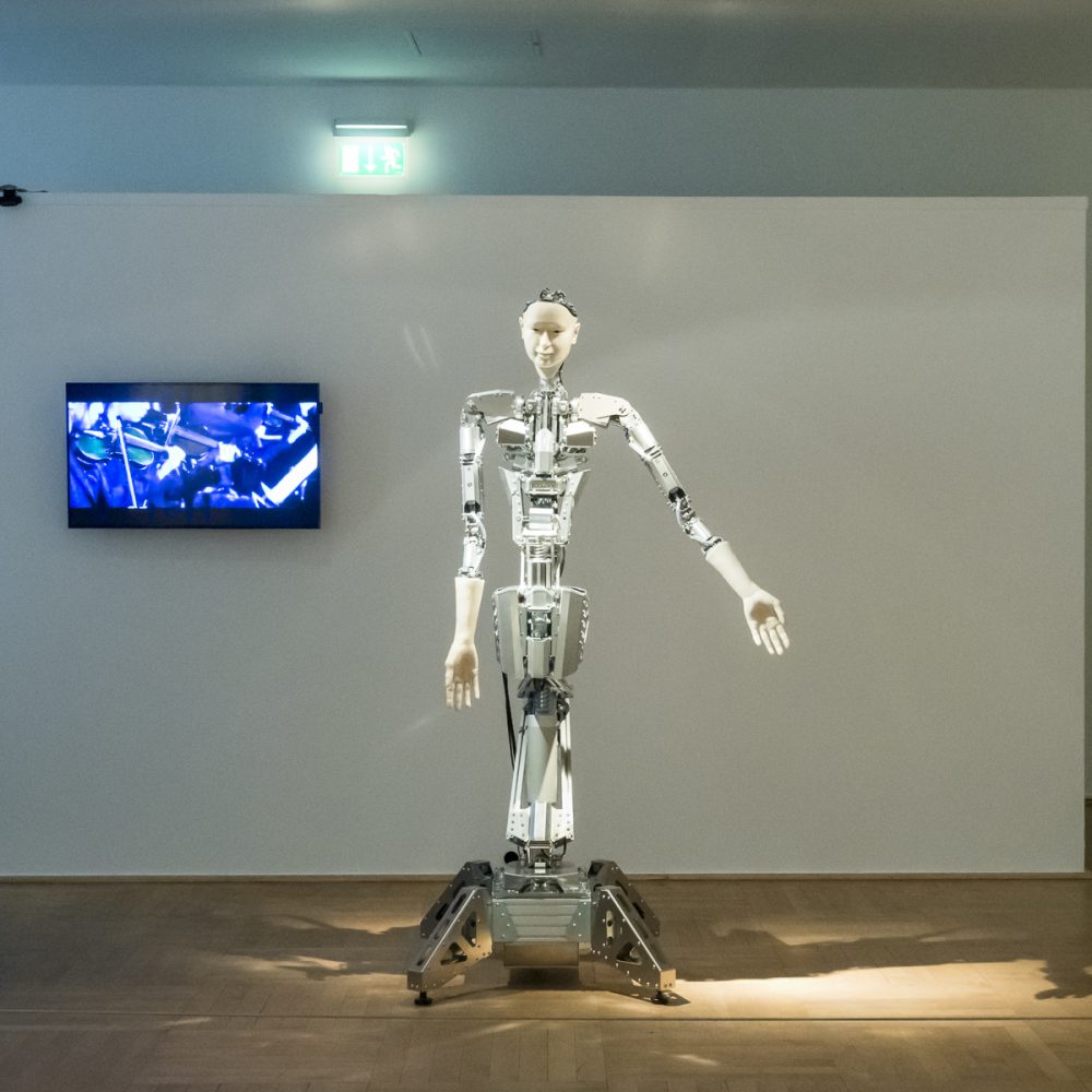 In the exhibition Körperwende, a robot stands in front of a white wall with a screen hanging on it