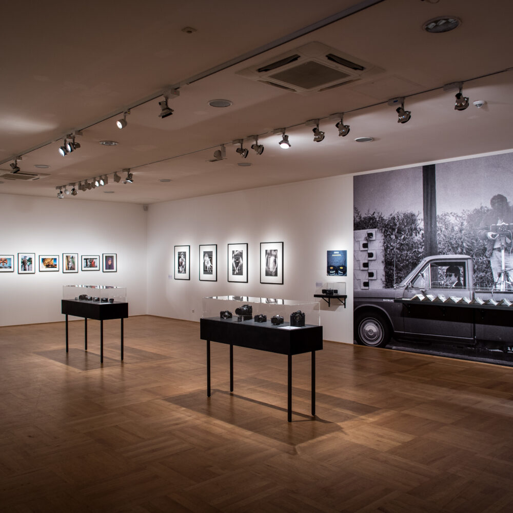 Ausstellungsansicht der Ausstellung 100 Jahre Nikon mit mehreren kleiner Fotografien, einer großen Fototapete sowie zwei schwarzen Vitrienen