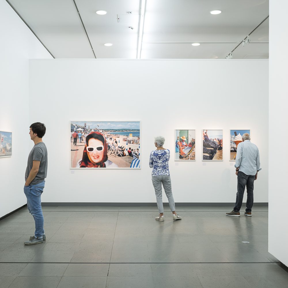 Mehrere Personen stehen in der Martin Parr Retrospektive und schauen sich Fotografien an