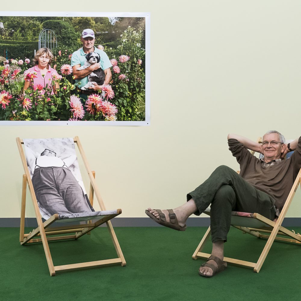 Der Fotograf Martin Parr sitzt auf einem Liegestuhl vor einer gelben Wand, an der ein Foto von ihm hängt in seiner Ausstellung