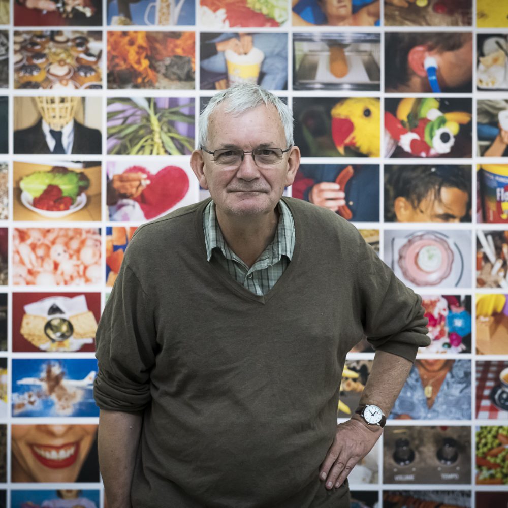 Martin Parr steht vor einer Wand an der viele bunte Aufnahmen von ihm hängen und beugt sich leicht nach vorne