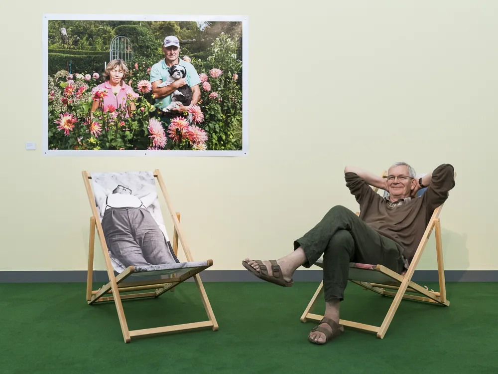 Photographer Martin Parr is sitting on a deck chair in front of a yellow wall with a photo of him hanging on it in his exhibition