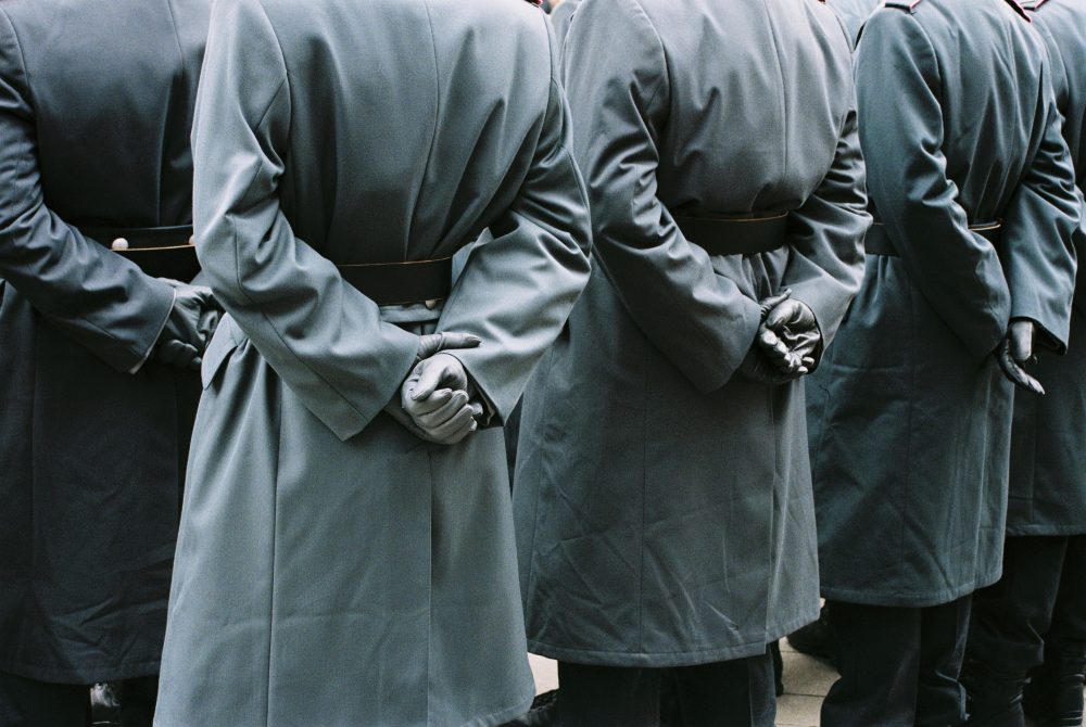 Eine Reihe von Personen in grauen Mänteln steht mit hinter dem Rücken verschränkten Händen, einige tragen schwarze Handschuhe.