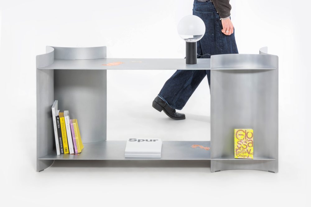 Eine Person geht an einem silbernen Regal mit Büchern, einer Lampe und einem gelben Buch darauf vorbei.