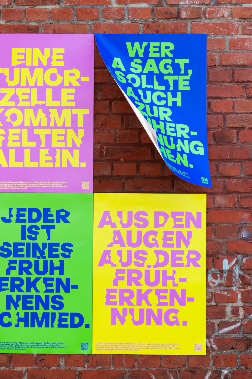 Vier plakative deutschsprachige Plakate in leuchtendem Pink, Blau, Grün und Gelb an einer roten Backsteinwand.