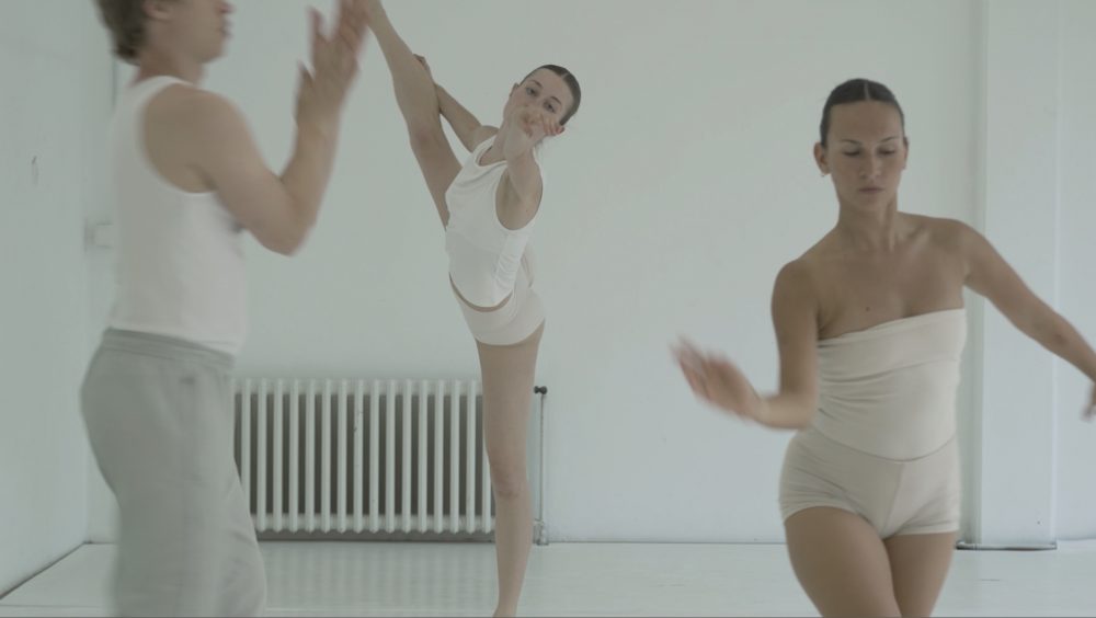 Drei Tänzerinnen in weißen Kleidern treten in einem hellen Studio auf; eine hebt ihr Bein hoch, während sich die anderen anmutig bewegen.