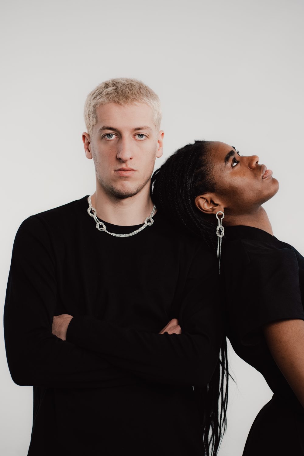 Ein Mann mit verschränkten Armen steht neben einer Frau, die sich auf seine Schulter stützt; beide tragen schwarze Kleidung und Silberschmuck.