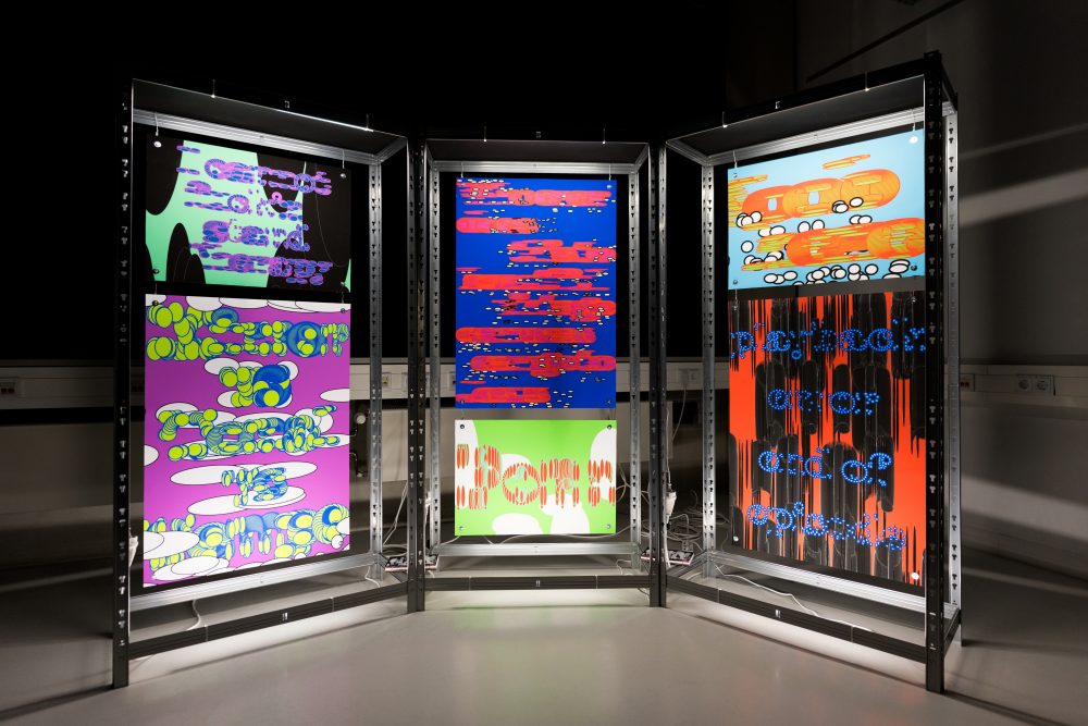 Drei Metallgestelle mit bunten, abstrakten digitalen Grafiken und Texten, hell beleuchtet in einem dunklen Raum.