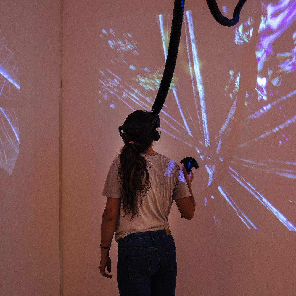 Woman standing in the Pendoran Vinci exhibition with VR glasses on her head and a controller in her hand, illuminated by projectors