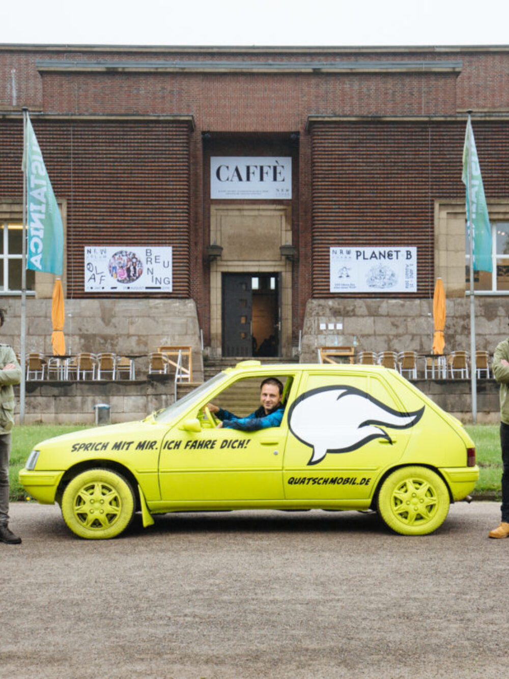 A yellow car in which a man is sitting stands in front of the NRW Forum. On the car it says: Talk to me. I will drive you