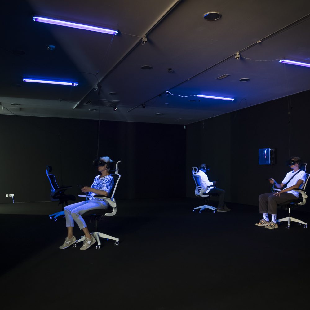 Drei Personen sitzen in der Ausstellung Whiteout auf Bürostühlen und haben eine VR Brille auf und werden dabei von Schwarzlicht angeleuchtet